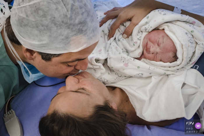 Photo by: <a href="https://www.fpja.com/documentary-family-photography-contests/clara-sampaio/rio-de-janeiro/2104416">Clara Sampaio - Brazil</a> Rio de Janeiro Brazil mom and dad kissing while holding their new baby - A triangle of much love