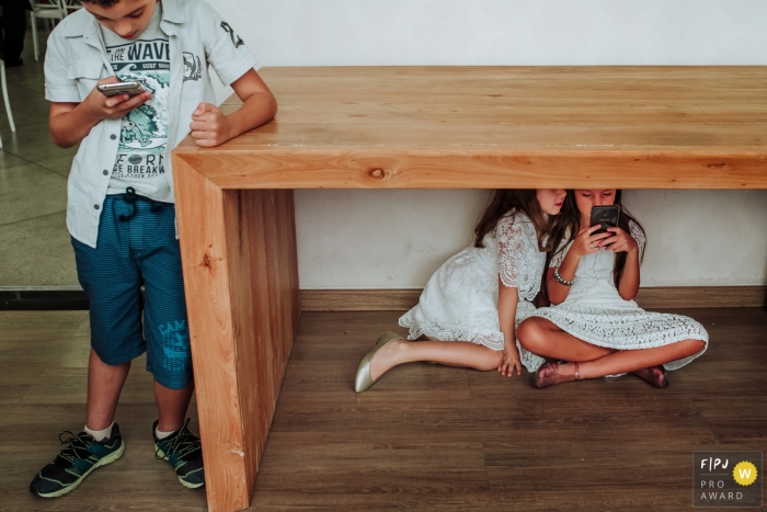 Photo by: <a href="http://fpja.com/documentary-family-photography-contests/sheila-maria-cupertino-gomes/minas-gerais/2102907">Sheila Maria Cupertino Gomes - Brazil</a> Minas Gerais Family Photography | Two girls under a table on a mobile device, with boy off the side also with a phone.