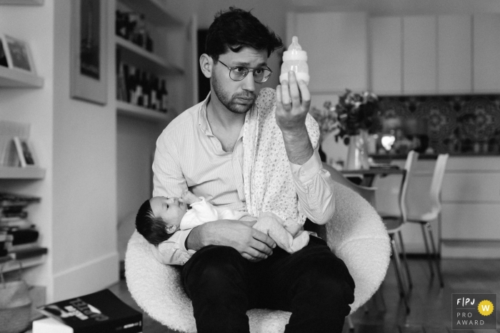 Photo by: <a href="https://www.fpja.com/documentary-family-photography-contests/marine-poron/paris/2103356">Marine Poron - France</a> Paris Ile-de-France family documentary report in Paris - dad holds up baby bottle to check levels