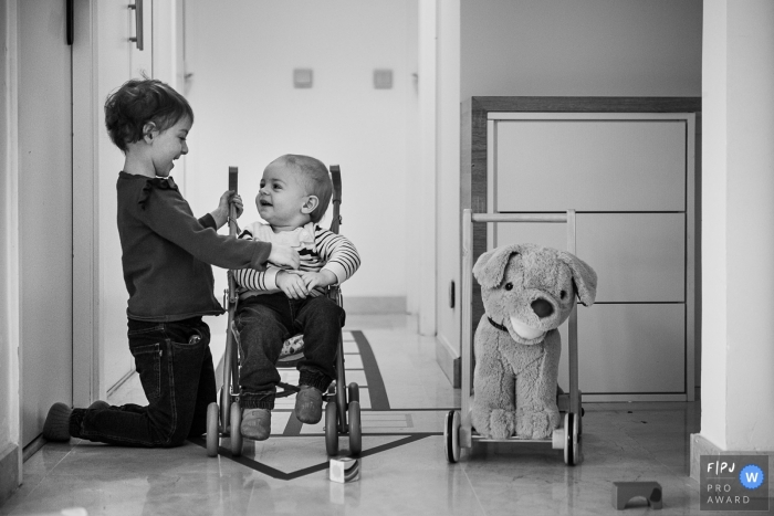 Photo by: <a href="https://www.fpja.com/documentary-family-photography-contests/nadine-court/savoie/2104002">Nadine Court - France</a> A little girl makes her little brother laugh by telling him stories while sitting in his stroller in France.