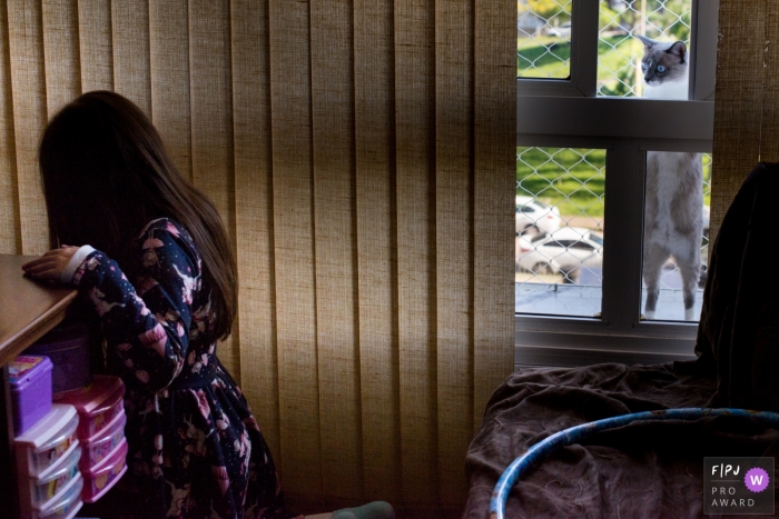 Photo by: <a href="https://www.fpja.com/documentary-family-photography-contests/morgana-secco/london/2104011">Morgana Secco - United Kingdom</a> London family image of a cat looking through the window to a girl in her bedroom.