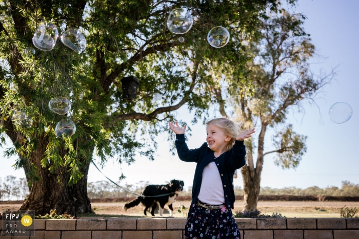 Photo by: <a href="https://www.fpja.com/documentary-family-photography-contests/linda-mauch/kent/2104306">Linda Mauch - United Kingdom</a> Kent England Little girl surrounded by bubbles outside with her dog
