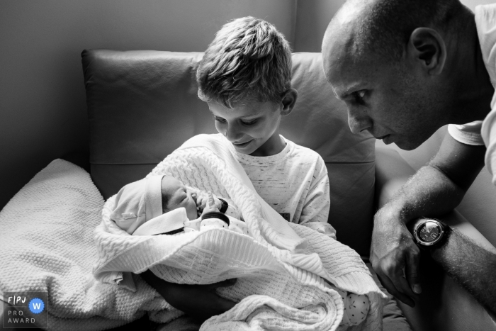 Photo by: <a href="https://www.fpja.com/documentary-family-photography-contests/thiago-braga/florianopolis/2106570">Thiago Braga - Brazil</a> Brother receiving the newborn with his father beside him admiring his son | Ilha Hospital e Maternidade family photo