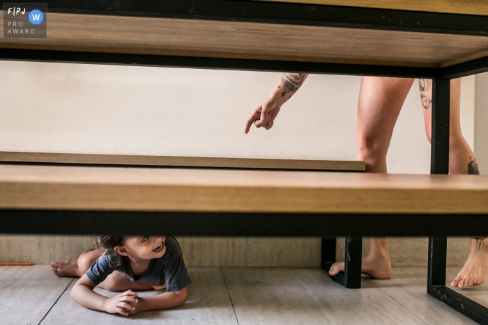 Photo by: <a href="https://www.fpja.com/documentary-family-photography-contests/adriana-costa/belo-horizonte/2105417">Adriana Costa - Brazil</a> Minas Gerais family photography of a daughter hiding from mother under the table.