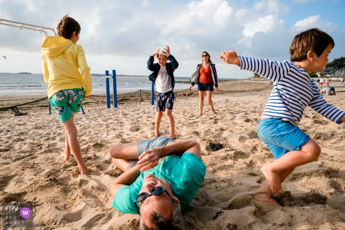 Photo by: <a href="https://www.fpja.com/documentary-family-photography-contests/sybil-rondeau/nantes/2105469">Sybil Rondeau - France</a> A dad rolls in the sand as the children run around during a family photo shoot