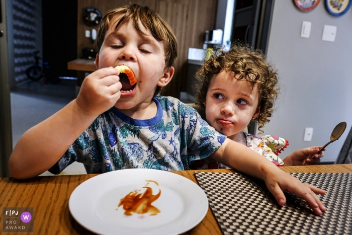 A young boy enjoys a snack at the table as his sibling watches intently | Minas Gerais family and kids photography