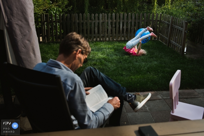 Photo by: <a href="https://www.fpja.com/documentary-family-photography-contests/charlene-van-der-gracht/amsterdam/2105973">Charlene van der Gracht - Netherlands</a> Young child plays on the grass in the yard as dad takes in a good book | Amsterdam Family Photographer