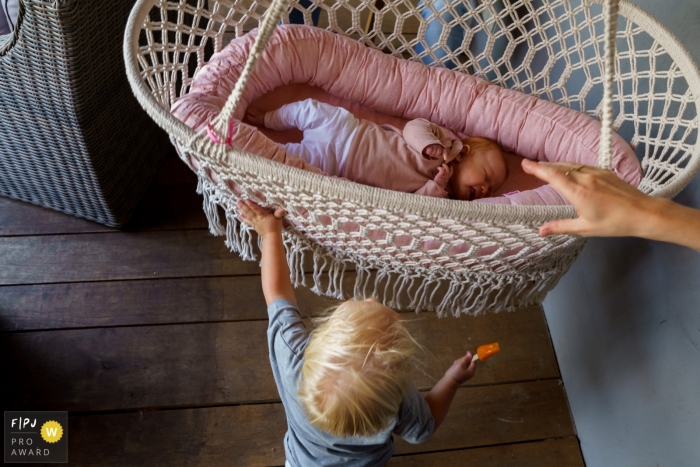 Photo by: <a href="https://www.fpja.com/documentary-family-photography-contests/indra-simons/overijssel/2106165">Indra Simons - Netherlands</a> Mom holds a hanging bassinet as a young sibling tries to swing his sister