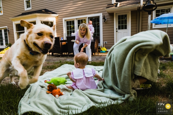 Photo by: <a href="https://www.fpja.com/documentary-family-photography-contests/pamela-anticole/pittsburgh/2106505">Pamela Anticole - United States</a> An image of backyard mayhem as dog runs off taking baby toys and big brother plays under blanket