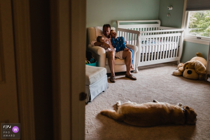 Photo by: <a href="https://www.fpja.com/documentary-family-photography-contests/pamela-anticole/pittsburgh/2106506">Pamela Anticole - United States</a> Pennsylvania family image of dad taking a moment in nursery with baby and son, while dog and stuffed dog lay around them
