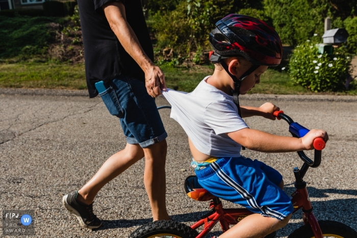 Photo by: <a href="https://www.fpja.com/documentary-family-photography-contests/pamela-anticole/pittsburgh/2106507">Pamela Anticole - United States</a> mom holds little boy's shirt as he rides his bike down the street during a family photo shoot in Pennsylvania