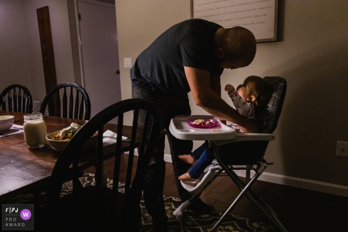 baby girl looks up at and reaches up for her dad as he buckles her into her high chair in their PA home.