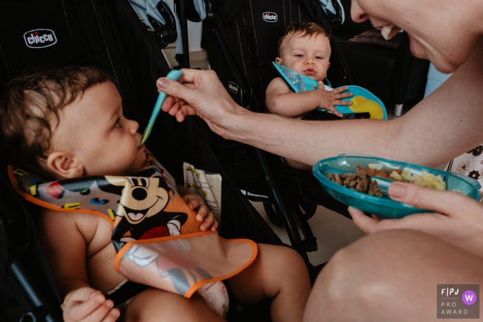 Photo by: <a href="https://www.fpja.com/documentary-family-photography-contests/thiago-braga/florianopolis/2106564">Thiago Braga - Brazil</a> Picture of a mother giving food to her two children, twin babies who are sitting in a car seat, waiting for the food.