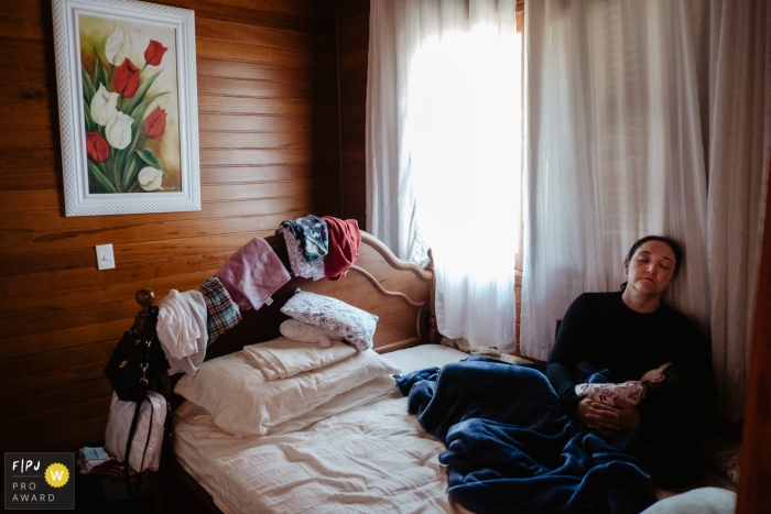 Mother, tired, with her daughter sleeping in a bedroom, with window light, on a Saturday afternoon. Clothes at the bottom, on top of the bed and headboard during a family photojournalism shoot in Brazil.