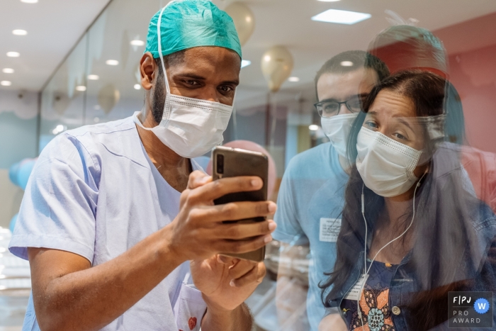 Photo by: <a href="https://www.fpja.com/documentary-family-photography-contests/claudia-ruiz/rio-de-janeiro/2108108">Claudia Ruiz - Brazil</a> Perinatal Laranjeiras Documentary Family Photo | father shows baby picture to family in nursery