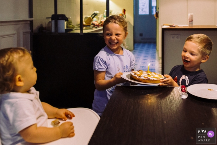 Photo by: <a href="https://www.fpja.com/documentary-family-photography-contests/marine-poron/paris/2107565">Marine Poron - France</a> Paris Documentary Family photo of siblings arriving at the table with a cake and one single candle to their younger brother