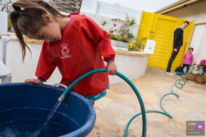 Photo by: <a href="https://www.fpja.com/documentary-family-photography-contests/annie-bang/los-angeles/2108226">Annie Bang - United States</a> Los Angeles Documentary Family Photographer | A girl fills up a bucket with water while her dad and her sister awaits.