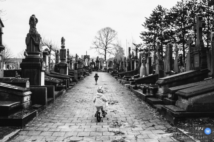 Photo by: <a href="https://www.fpja.com/documentary-family-photography-contests/christophe-de-mulder/antwerpen/2108875">Christophe De Mulder - Belgium</a> Black and white Antwerpen documentary-style family photography session showing a brother and sister biking through the graveyard in Belgium