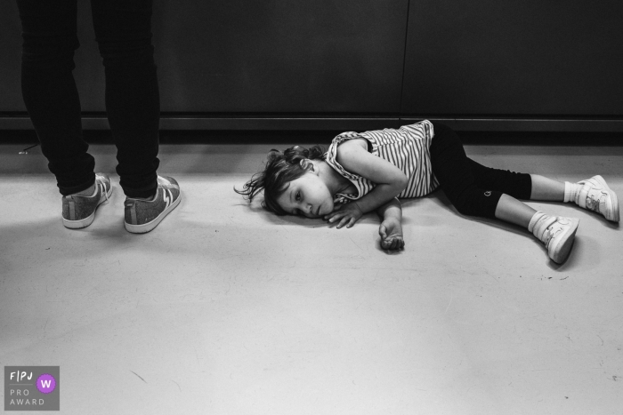 Photo by: <a href="https://www.fpja.com/documentary-family-photography-contests/christophe-de-mulder/antwerpen/2108878">Christophe De Mulder - Belgium</a> Flanders family photography session image of a girl lying on the ground at the cash register of another shop mom wanted to visit