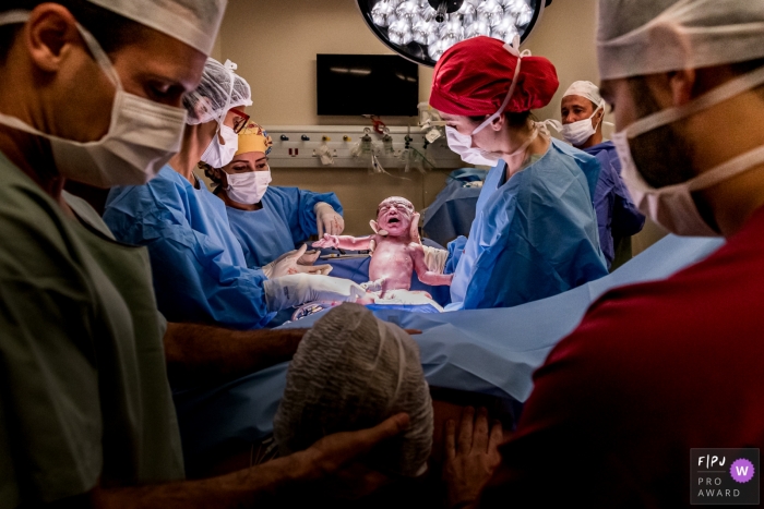 Photo by: <a href="https://www.fpja.com/documentary-family-photography-contests/liliane-cotta/rio-de-janeiro/2111483">Liliane Cotta - Brazil</a> Complexo Hospitalar de Niterói birth picture in a documentary-style of the Obstetrician showing parents their baby that was just born while the whole team is focused on the baby the anesthesiologist is the only one who is looking at the mother
