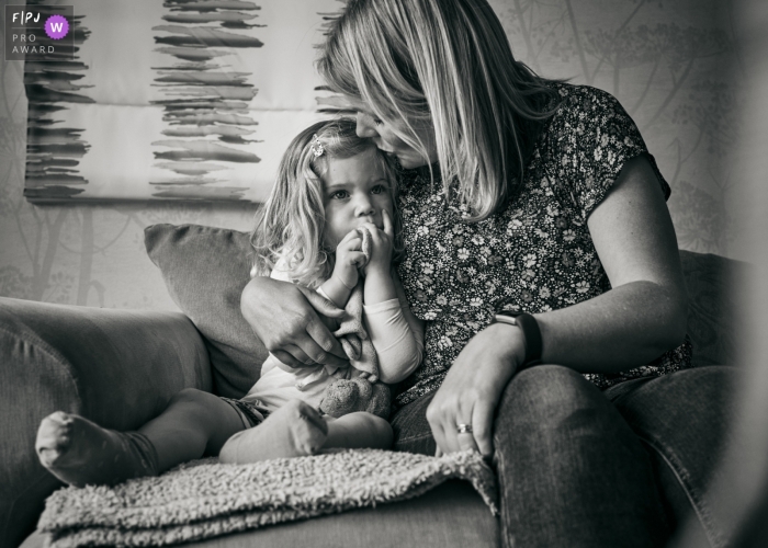 Day in the Life photography session at home in England showing A mother kisses her daughter as she snuggles in Day in the Life photography session at home in England showing A mother kisses her daughter as she snuggles in