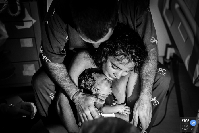 Photographie de naissance de l'hôpital Plantadores de Cana, animée par le moment, recouverte de noir et blanc d'une mère et d'un père tenant leur nouveau-né