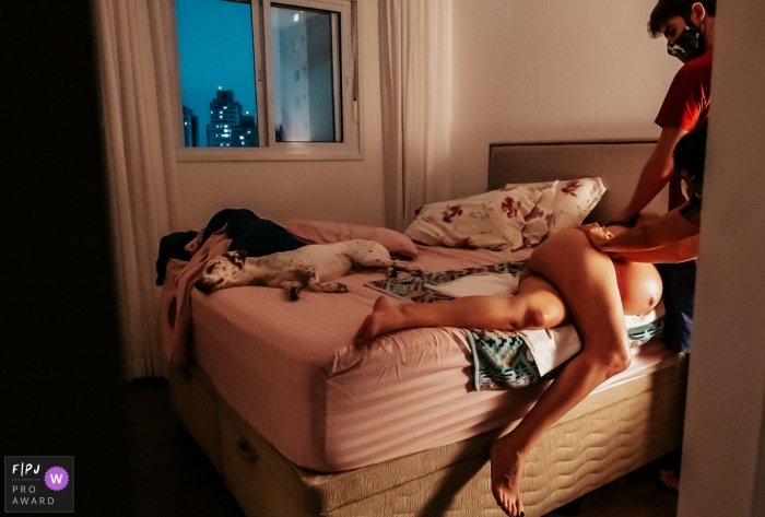 Photographie de naissance à domicile à Sao Paulo d'une femme allongée sur un lit avec vue sur les gratte-ciel de la ville par la fenêtre Photographie de naissance à domicile à Sao Paulo d'une femme allongée sur un lit avec vue sur les gratte-ciel de la ville par la fenêtre