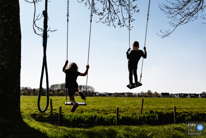 Moment driven Groningen family photojournalism image of a brother and sister standing on the seats of swings and they soar Moment driven Groningen family photojournalism image of a brother and sister standing on the seats of swings and they soar