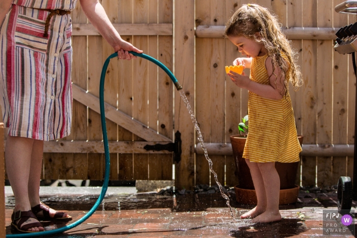 Moment driven New Hampshire family photojournalism image of Mom holds hose for young daughter to fill her cup on a hot day Moment driven New Hampshire family photojournalism image of Mom holds hose for young daughter to fill her cup on a hot day