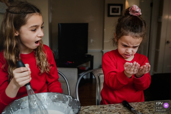 Moment-driven New Hampshire family photography of sisters making pancakes react to messy batter on little sister's hands