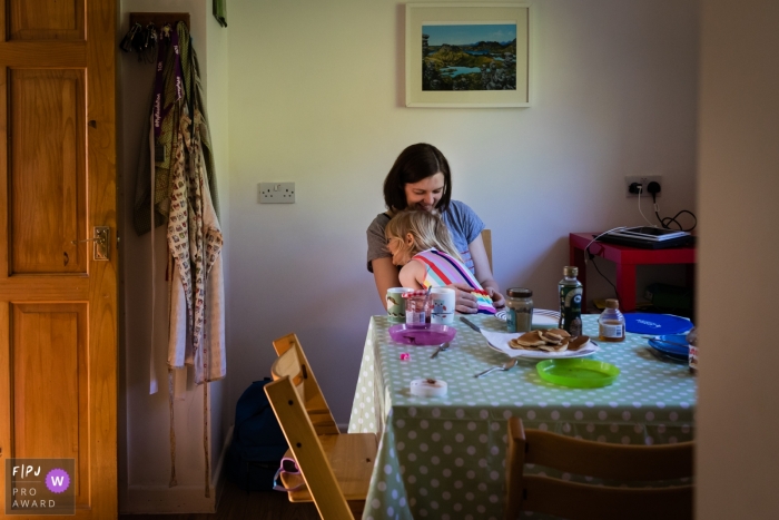 Image de photojournalisme familial du Cambridgeshire animée par un moment capturant la mère et la fille souriant tout en se faisant un câlin à la table du petit-déjeuner à la maison