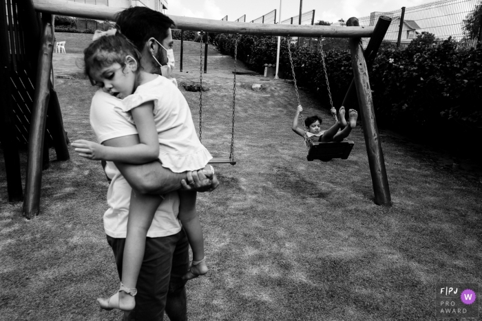 Image de photojournalisme de la famille Florianopolis animée par un moment en BW montrant un père avec sa fille dormant sur ses genoux tandis que l'autre fils joue de la balançoire