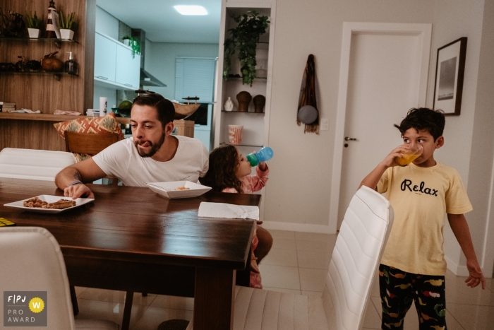 Photographie de famille de Florianopolis animée par un moment montrant une famille en train de dîner, avec un père montrant sa langue à cause de la faim, un fils buvant du jus et une fille à côté de lui juste après avoir mangé