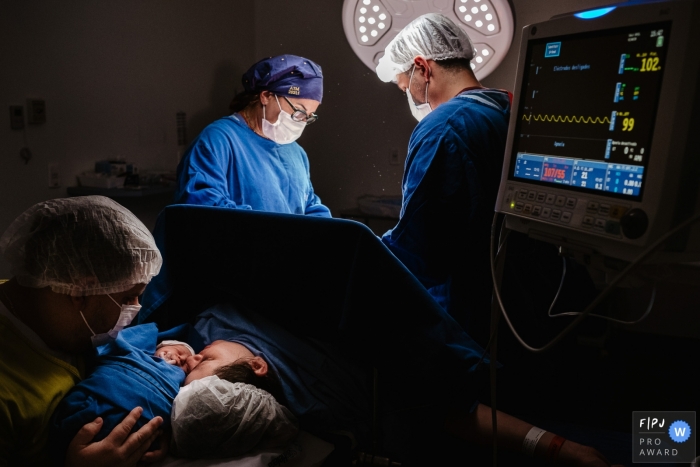 Photo de naissance de Maternidade Santa Helena montrant le moment juste après la césarienne avec l'équipe médicale en train de finir et les parents étreignant leur fille