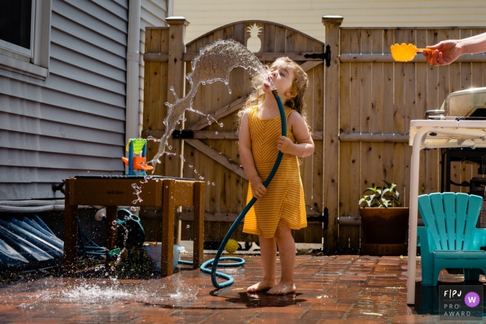 Image de famille d'arrière-cour du New Hampshire d'une jeune fille prenant un verre dans un tuyau d'arrosage alors que maman tient une tasse