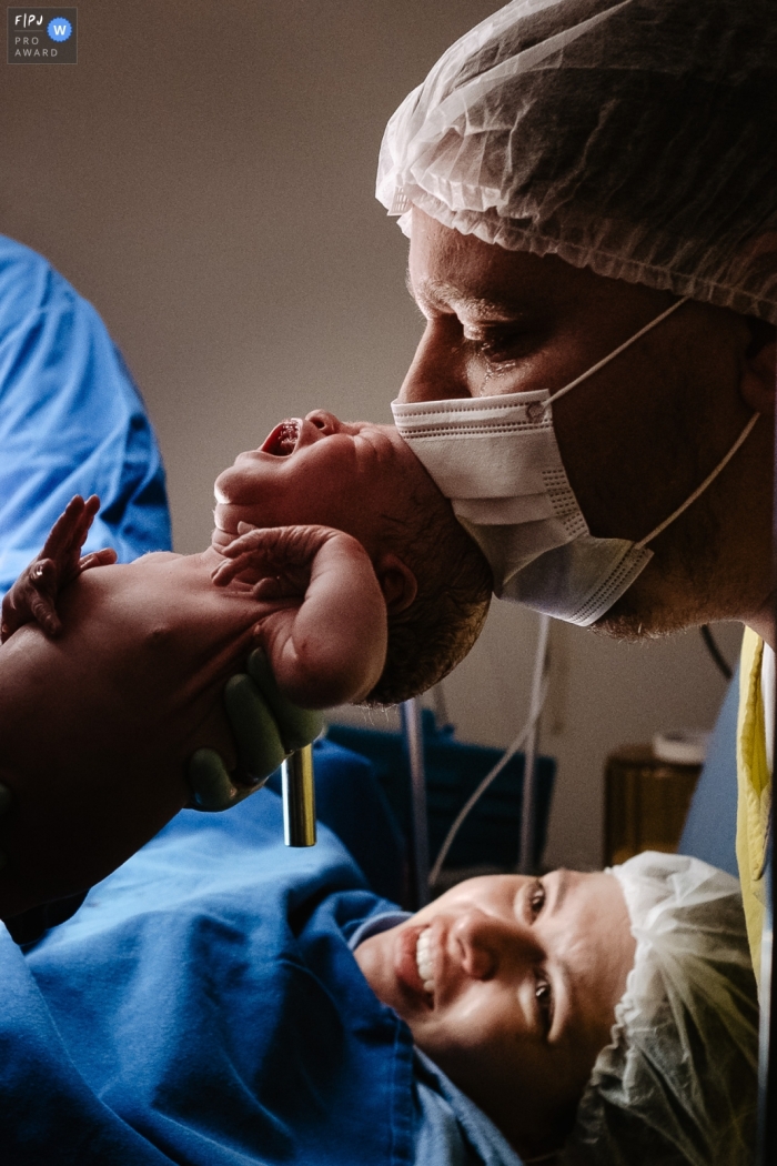 Photographie documentaire de la naissance de Florianpolis à la Maternidade Santa Helena faisant la chronique du père émotif et pleurant juste après la première rencontre avec sa fille qui vient de naître avec la mère en arrière-plan regardant avec admiration