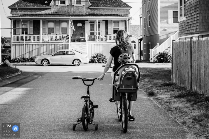 New Hampshire dokumentarische Familienfotografie einer Mutter, die Fahrrad fährt, mit einem kleinen Jungen im Tragesitz, während sie sich an seinem verlassenen Fahrrad festhält, während sie nach Hause fahren