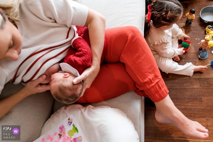 Dokumentarische Familienfotografie aus Sao Paulo, die eine Mutter zeigt, die zu Hause stillt, während ein Kleinkind mit Spielzeug auf dem Boden spielt