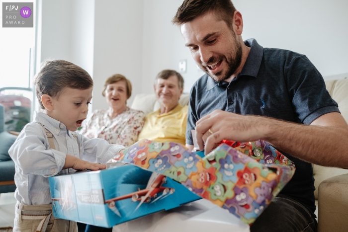 Florianopolis, Brasilien Familienfotografie eines Kindes, glücklich und überrascht, sein Geburtstagsgeschenk öffnend, mit seinem Vater und seinen Großeltern im Hintergrund