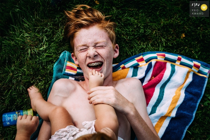 A New Hampshire documentary family photographer captured this image of a boy with braces laughing as baby sister puts foot on his face A New Hampshire documentary family photographer captured this image of a boy with braces laughing as baby sister puts foot on his face