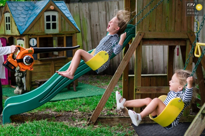 Mom takes a break from yard clean up to blow the leaf blower at her kids on their swing set during this outdoor Sarasota documentary family photography shoot