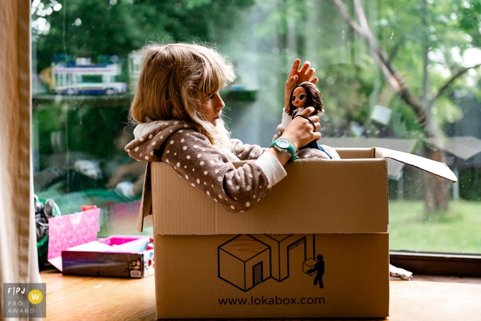 Documentary family photojournalism from Wallonie, BE with a girl sitting in a cardboard box by a window and playing with an lol doll Documentary family photojournalism from Wallonie, BE with a girl sitting in a cardboard box by a window and playing with an lol doll