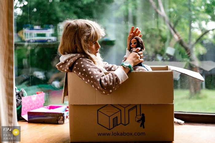 Documentary family photojournalism from Wallonie, BE with a girl sitting in a cardboard box by a window and playing with an lol doll