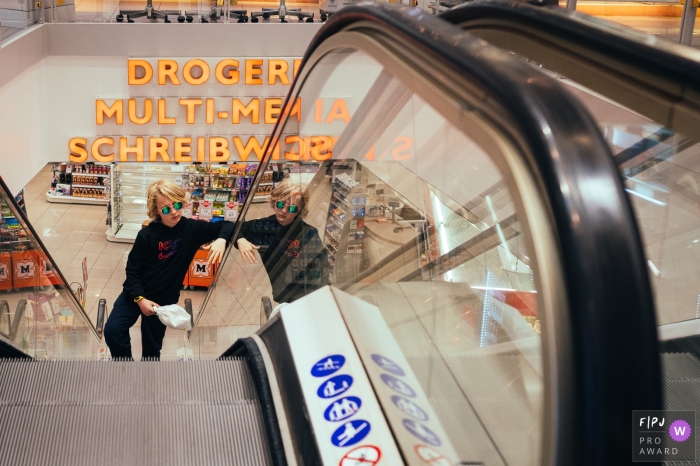 Photographie de style documentaire d'Amsterdam d'une séance familiale capturant un garçon dans une pharmacie sur un escalator, jouant cool