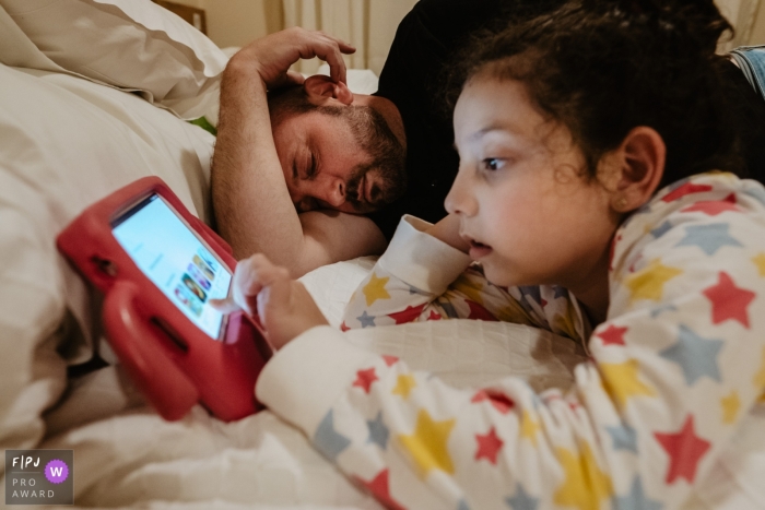 Photographie documentaire de la famille Florianopolis à Santa Catarina capturant un père et sa fille allongés dans leur lit à l'heure du coucher, une fille regardant une tablette et un père fatigué de dormir