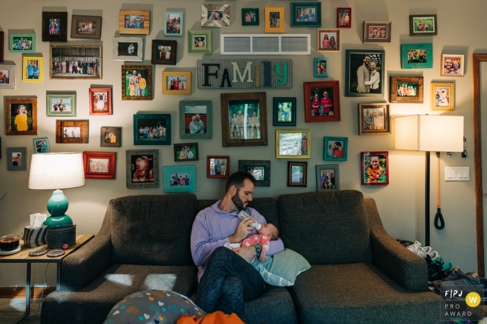 Vader zit op de bank terwijl hij zijn pasgeborene voedt terwijl hij wordt omringd door een enorme familiewand in hun huis in Madison, Wisconsin